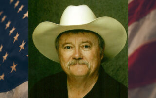 Photo of John Compere wearing a white cowboy hat, against an American flag background