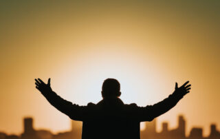 Silhouetted man standing with hands raised, overlooking sunset-lit city skyline
