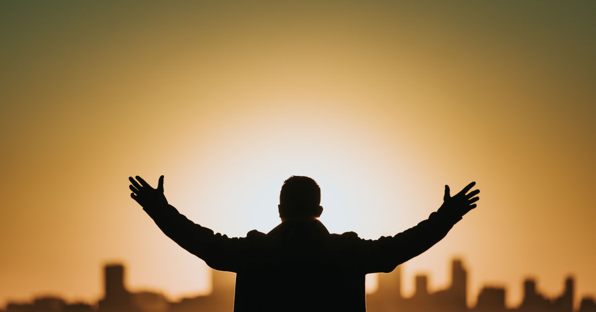Silhouetted man standing with hands raised, overlooking sunset-lit city skyline