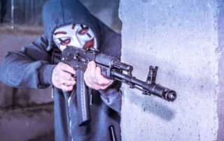 Disturbing image of masked, hooded person standing behind concrete wall, pointing a black rifle