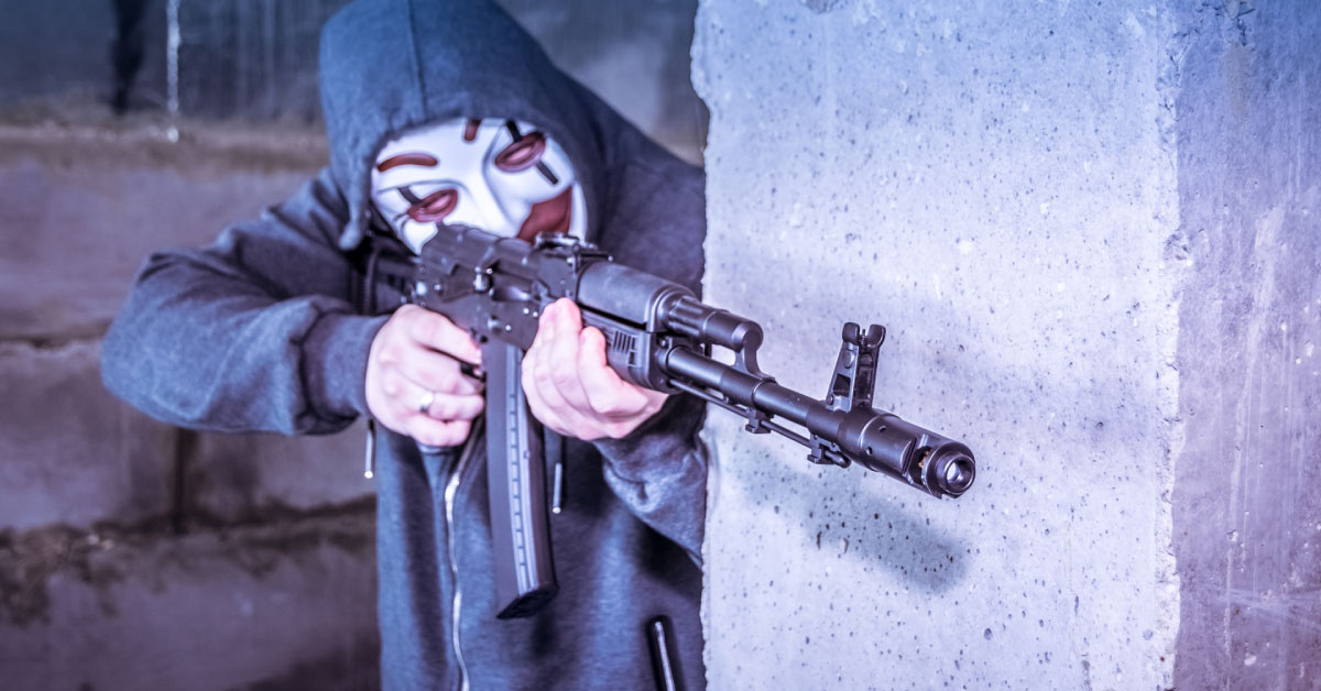 Disturbing image of masked, hooded person standing behind concrete wall, pointing a black rifle