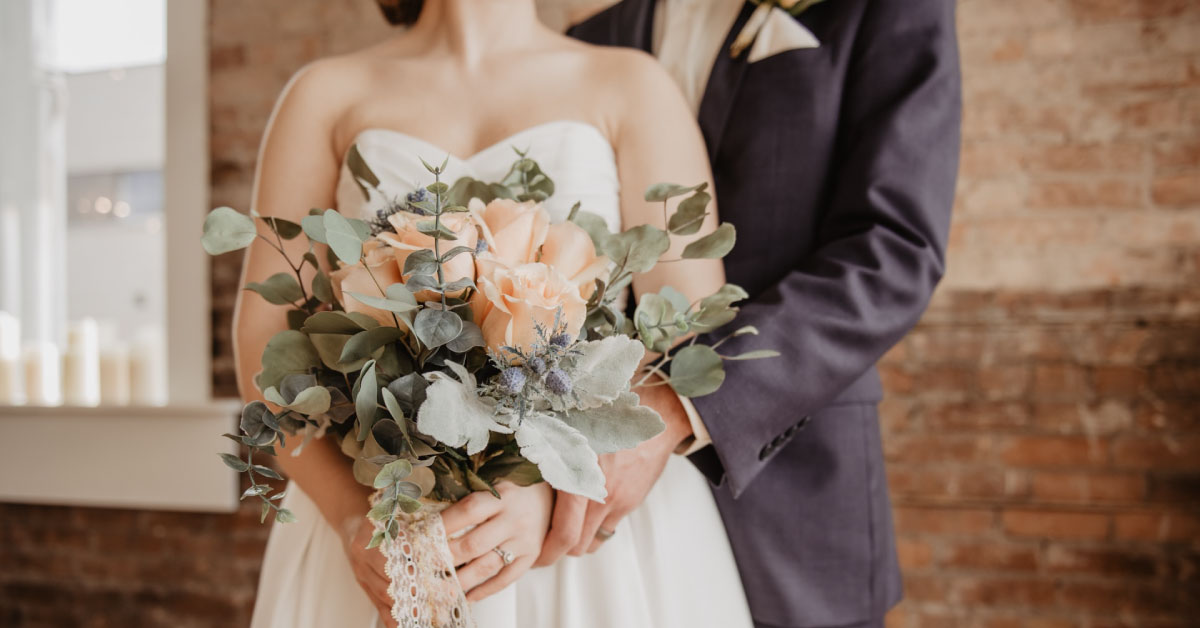 Woman wearing wedding dress holding flowers, standing in front of man wearing suit