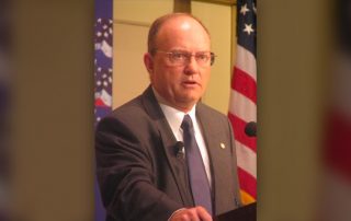 Colonel Lawrence Wilkerson at a lectern with American flags behind.