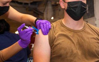 Hospital Corpsman with purple gloves administering COVID vaccine shot to another US servicemember.