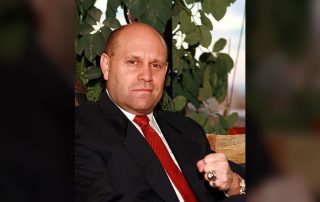 Photo of Mikey Weinstein looking intently toward the camera with a slightly raised fist, wearing a black suit and red tie.