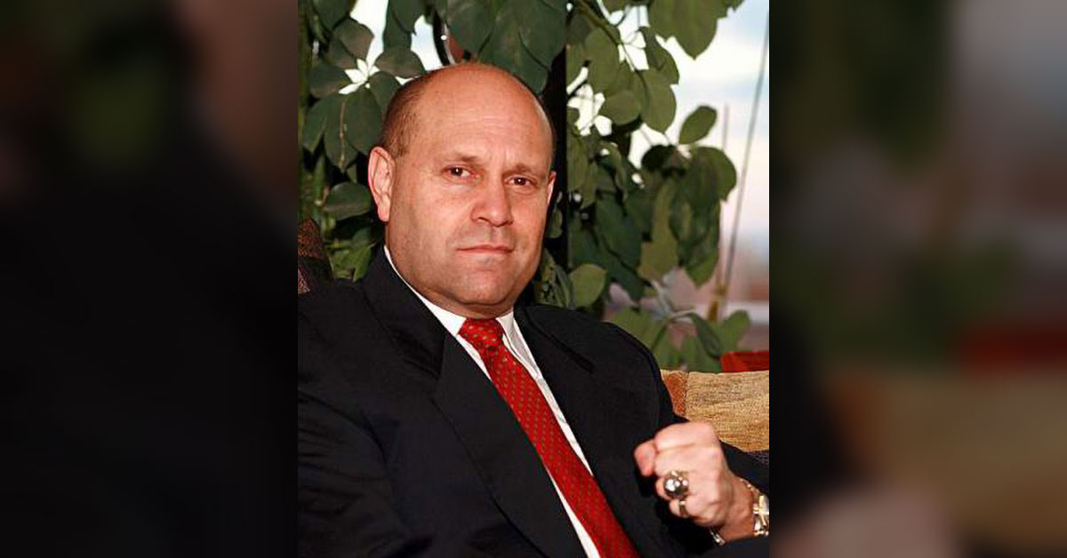 Photo of Mikey Weinstein looking intently toward the camera with a slightly raised fist, wearing a black suit and red tie.
