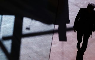 View from above of a man obscured in shadow walking on a stone floor with larger shadows of building structure around him.