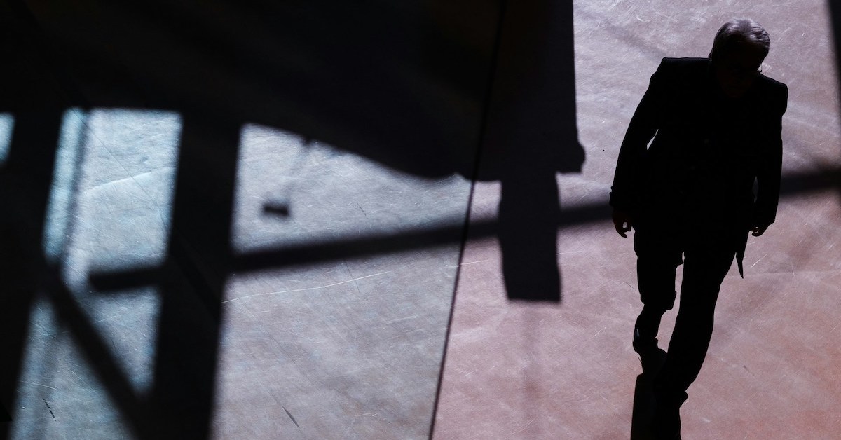 View from above of a man obscured in shadow walking on a stone floor with larger shadows of building structure around him.