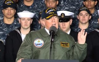 Trump when he was president dressed in navy jacket and hat speaking with group of sailors behind him
